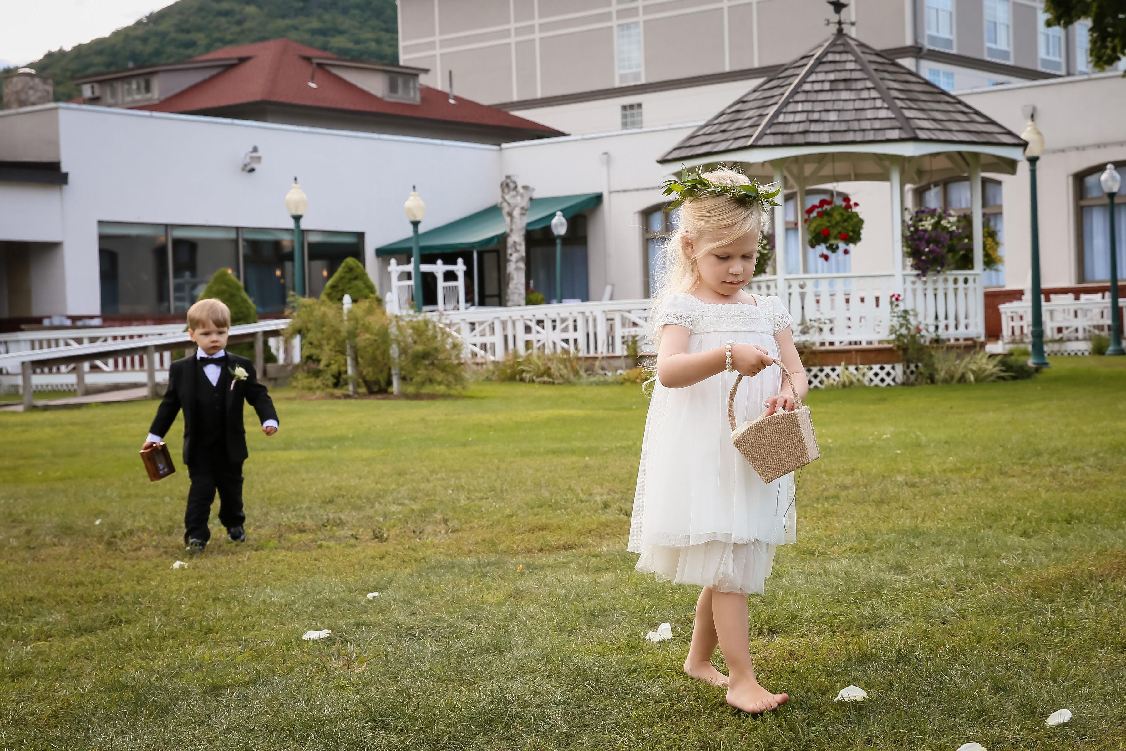 Toddler flower girl walks across the lawn with toddler ring bearer following