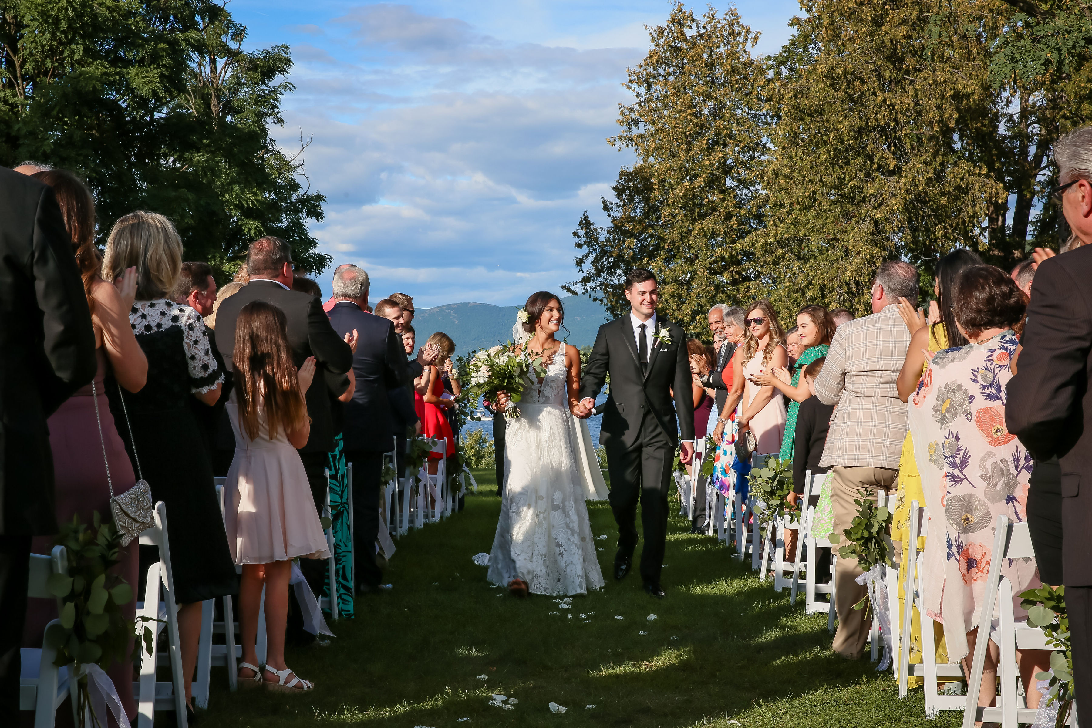 Happy newlyweds walk down the aisle as guests stand to either side. Mountains in the background